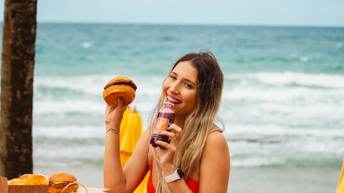 Smiling woman holding a burger and drink while enjoying a day at the beach.
